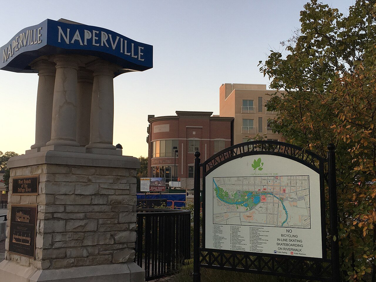 Stone Naperville Riverwalk entrance sign and visitor map beside autumn trees on Water Street.